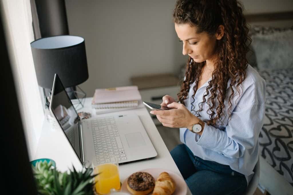A woman at a desk looking at her smartphone with a laptop open beside her