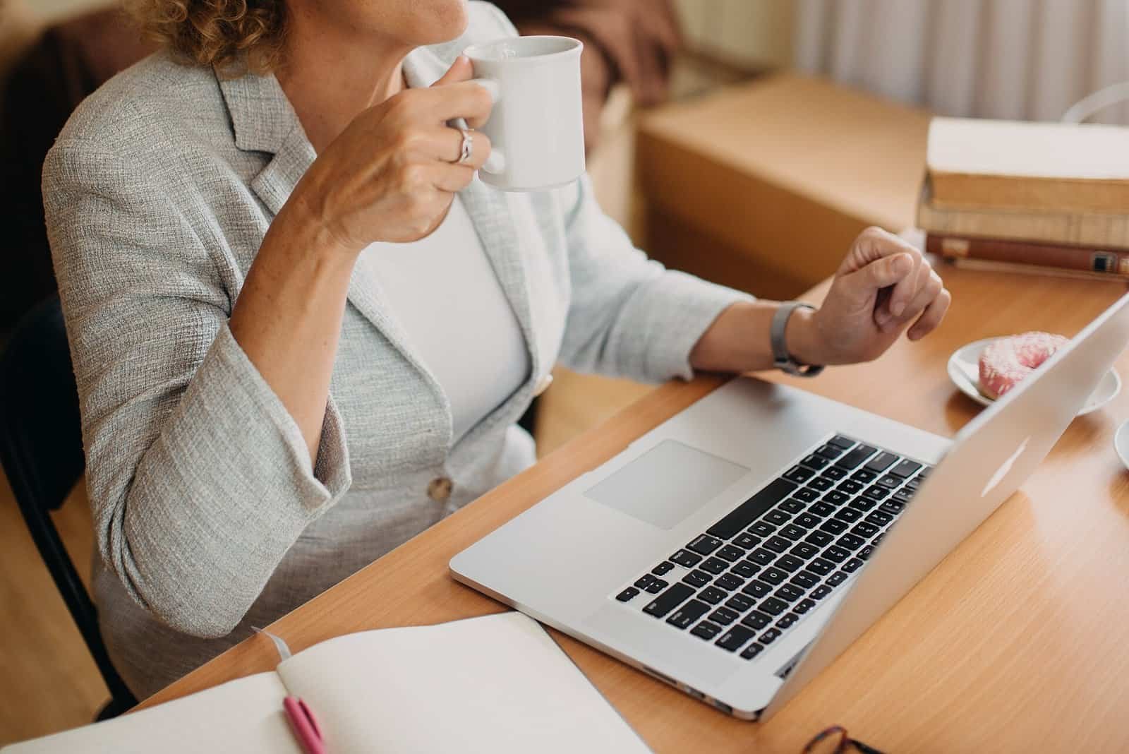 A person holding a coffee cup while looking at a laptop on a wooden desk