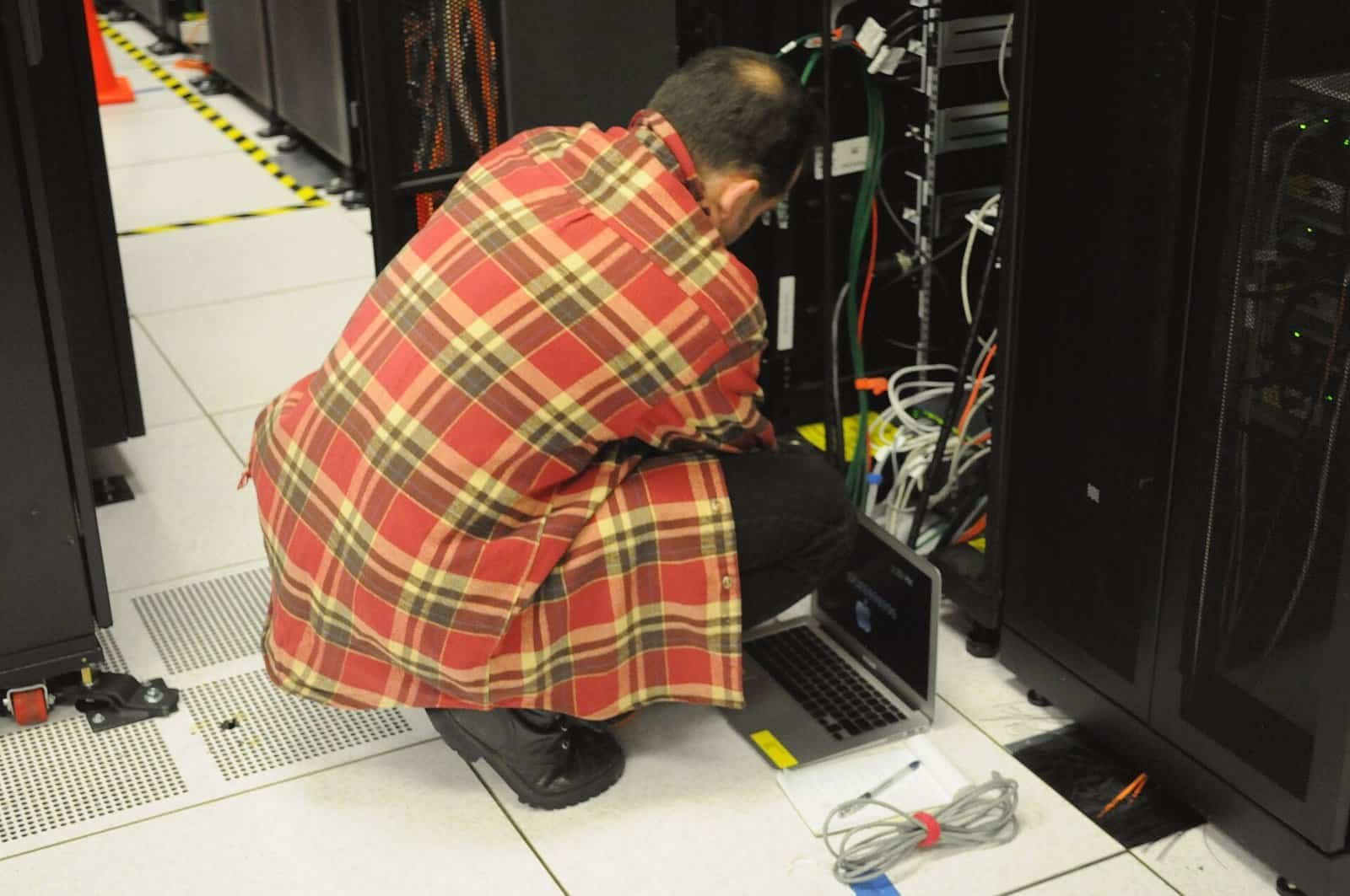 A technician crouching to work on a data center server rack with a laptop